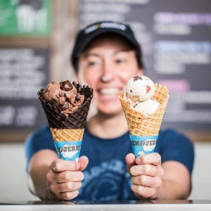 A staff member at Ben and Jerry's holding two waffle cones full of ice cream, one dipped in chocolate and one that is not.